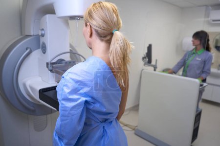 Foto de Paciente femenina de pie frente a la máquina de mamografía operada por mamógrafo mediante consola de control - Imagen libre de derechos