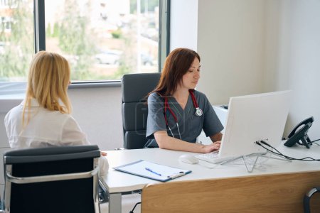 Téléchargez les photos : Médecin assis au bureau en tapant sur PC de bureau en présence du client pendant la consultation - en image libre de droit