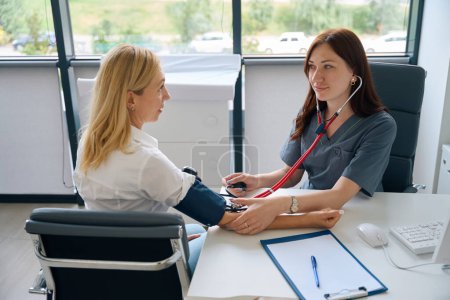 Foto de Médico general sentado en el escritorio midiendo la presión arterial del paciente femenino usando con esfigmomanómetro - Imagen libre de derechos