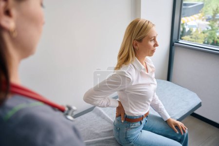 Photo for Female patient seated on examination couch touching her lower back in presence of doctor - Royalty Free Image