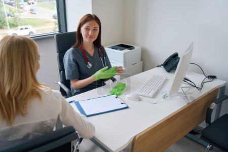 Photo for Calm physician seated at desk in office talking to patient while taking off gloves after medical checkup - Royalty Free Image