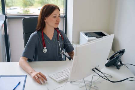 Photo for Waist-up portrait of focused physician with stethoscope around her neck working on desktop PC - Royalty Free Image