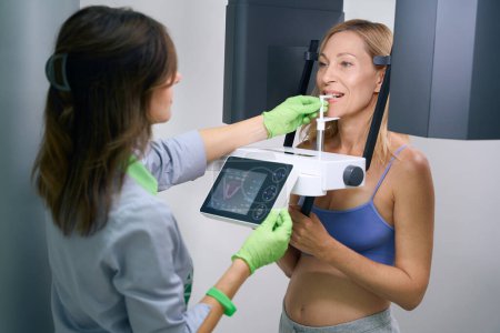 Téléchargez les photos : Radiographe insérant le bloc de morsure dans la bouche du patient féminin avant la radiographie panoramique numérique dentaire - en image libre de droit