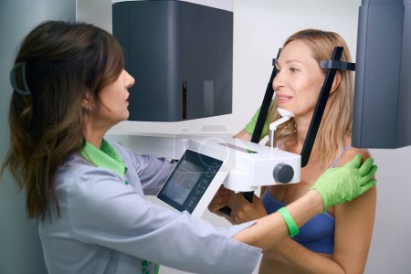 Téléchargez les photos : Technologue en radiologie expérimenté positionnant une patiente adulte à l'intérieur d'un scanner de tomodensitométrie à faisceau conique dentaire - en image libre de droit