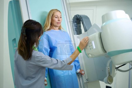 Foto de Paciente apoyado contra la mesa de examen radiográfico mientras el tecnólogo radiológico ajusta el ángulo de inclinación del tubo de rayos X - Imagen libre de derechos
