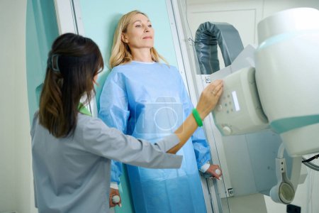 Foto de Mujer apoyando su espalda contra la mesa de examen radiográfico mientras el radiografo ajusta el ángulo de inclinación del tubo de rayos X - Imagen libre de derechos