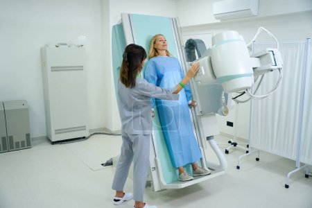 Foto de Mujer apoyando la espalda contra la mesa radiográfica mientras la tecnóloga radiológica ajusta el ángulo de inclinación del tubo de rayos X - Imagen libre de derechos