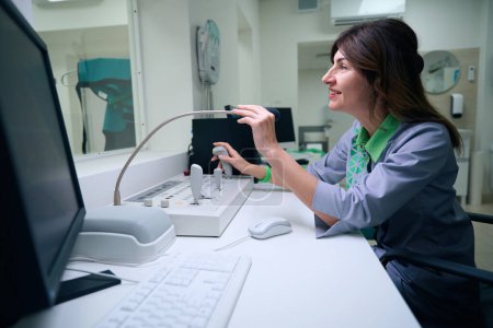 Photo for Side view of smiling radiographer seated at desk talking into microphone while moving joystick on operating console in control room - Royalty Free Image