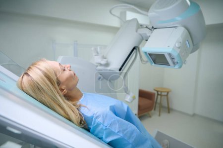 Foto de Vista lateral de una paciente vestida en posición supina sobre una mesa radiográfica inclinada hacia un tubo de rayos X - Imagen libre de derechos