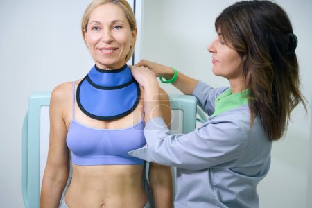 Photo for Smiling female patient leaning against radiographic bucky wall stand while radiographer wrapping thyroid collar around her neck - Royalty Free Image
