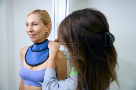 Foto de Alegre dama apoyada en soporte de pared bucky de rayos X mientras el tecnólogo radiológico envuelve el escudo tiroideo alrededor de su cuello - Imagen libre de derechos
