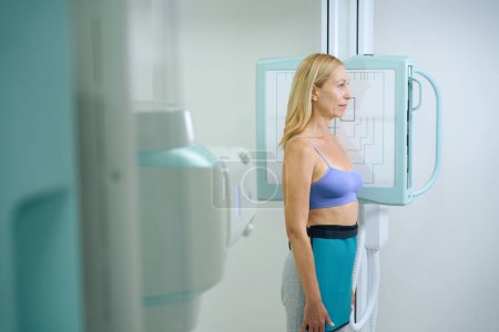 Téléchargez les photos : Patiente debout avec son côté gauche positionné contre le récepteur d'image devant la machine à rayons X - en image libre de droit