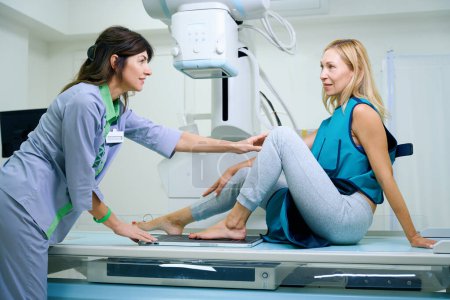 Foto de Paciente femenina sentada en mesa de rayos X mientras la tecnóloga radiológica coloca su pie izquierdo con superficie plantar en el receptor de imagen - Imagen libre de derechos