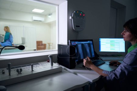 Radiógrafo enfocado sentado en computadoras de escritorio mirando a la mujer en la mesa radiográfica a través de la ventana en la sala de control