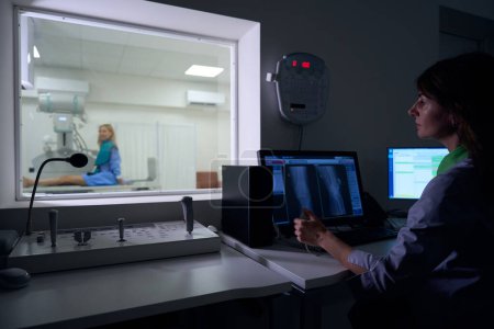 Foto de Tecnólogo radiológico serio sentado en computadoras de escritorio mirando al paciente en la mesa de rayos X a través de la ventana en la sala de control - Imagen libre de derechos