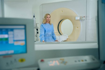 Téléchargez les photos : Tranquille dame en robe d'examen jetable assis sur la table CT dans la salle de scan - en image libre de droit