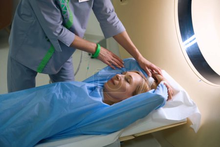 Foto de Paciente femenina acostada en la mesa de TC con los brazos elevados por encima de la cabeza ayudada por un radiografo - Imagen libre de derechos