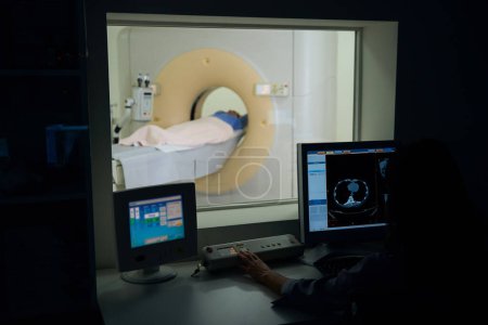 Foto de Tecnólogo radiológico sentado en el ordenador de sobremesa pulsando el botón de la consola de operación en la sala de control CT - Imagen libre de derechos