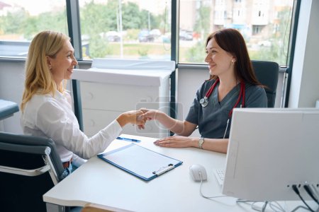 Photo for Physician seated at desk in her office shaking hands with female client after signing informed consent form - Royalty Free Image