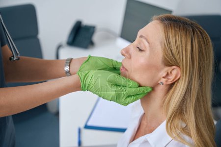 Téléchargez les photos : Otolaryngologue expérimenté dans les gants en nitrile palpant les glandes salivaires du patient pendant l'examen physique - en image libre de droit