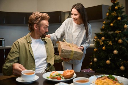 Foto de Mujer feliz dando regalo a hombre guapo mientras que tienen cena de Año Nuevo festiva celebrando vacaciones juntos - Imagen libre de derechos