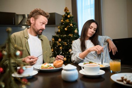Téléchargez les photos : Femme bouleversée assise à la table à la maison pendant la célébration de Noël avec le visage offensé dîner avec l'homme - en image libre de droit
