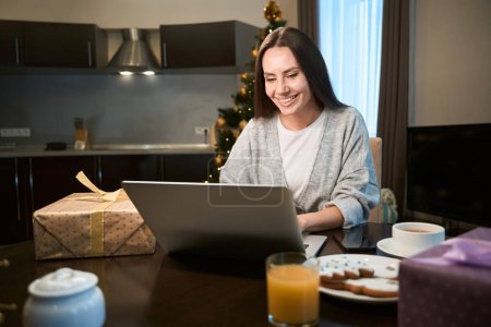 Foto de Mujer sonriente rodeada de regalos utilizando el ordenador portátil para las compras en línea en las vacaciones de Navidad en casa - Imagen libre de derechos