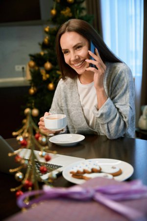 Foto de Mujer alegre disfrutando del café caliente mientras felicita a su amigo con las vacaciones de Navidad - Imagen libre de derechos