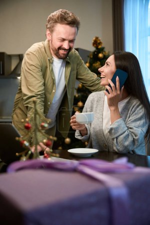 Téléchargez les photos : Joyeux homme et femme assis à la maison dans la chambre décorée en buvant du café ayant un appel téléphonique - en image libre de droit