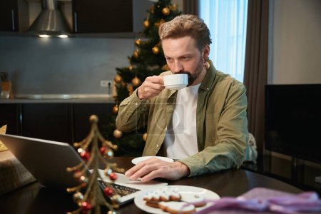Téléchargez les photos : Homme heureux faire appel vidéo dans la chambre avec arbre de Noël et profiter du café chaud pendant les vacances d'hiver - en image libre de droit