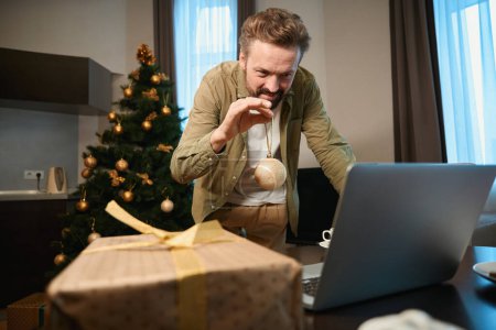 Photo for Attractive man talking online via laptop showing Christmas decorations to computer camera while preparing for holidays - Royalty Free Image
