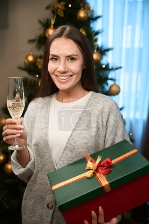 Photo for Beautiful woman toasting with glass of champagne holding wrapped present box at New Year party - Royalty Free Image