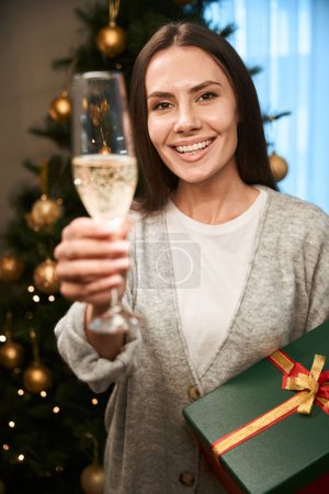 Photo for Joyful female raising glass of champagne with present box in hand celebrating Christmas holidays in hotel - Royalty Free Image