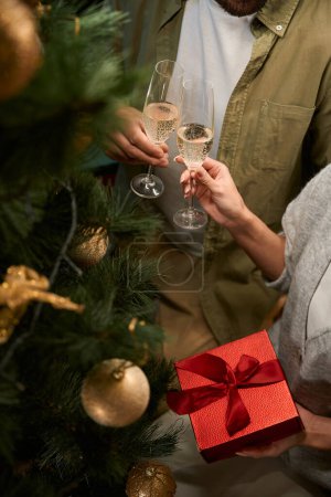 Foto de Mujer alegre con caja de regalo tostadas con copas de champán con el hombre mientras se celebra la Navidad - Imagen libre de derechos
