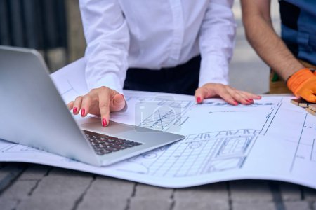 Photo for Cropped photo of female engineer using laptop and architectural drawings in presence of builder - Royalty Free Image