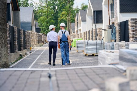 Photo for Back view of construction supervisor and contractor walking along street between rows of unfinished residential houses - Royalty Free Image