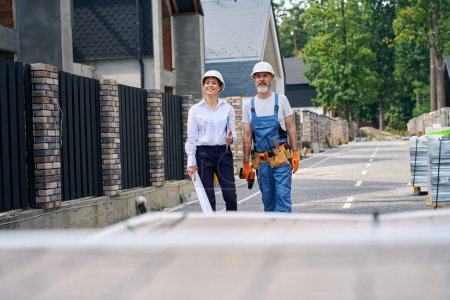 Téléchargez les photos : Superviseur de chantier souriant et travailleur de la construction debout sur la rue devant une maison résidentielle inachevée - en image libre de droit