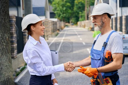 Photo for Side view of construction inspector and builder shaking hands on street among unfinished cottages - Royalty Free Image