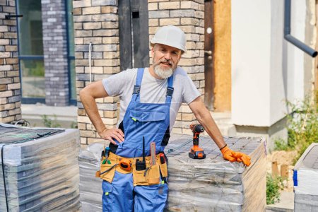 Photo for Smiling construction worker in hard hat leaning on stack of paving blocks placed in front of unfinished residential house - Royalty Free Image