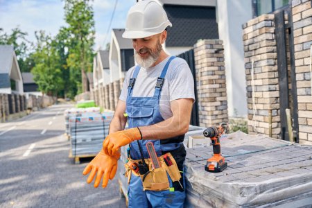 Photo for Pleased bricklayer in hard hat putting on rubber gloves while leaning against palletized paving blocks in middle of street - Royalty Free Image