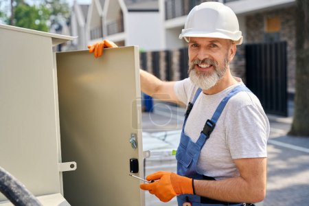 Photo for Waist-up portrait of smiling electrician with screwdriver in his hand leaning on power distribution box door outdoors - Royalty Free Image