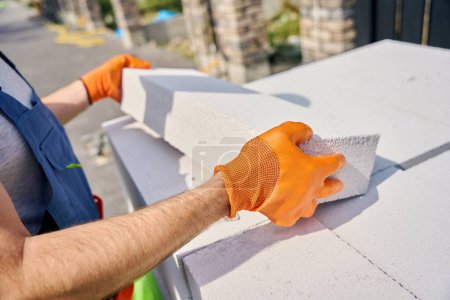 Foto de Foto recortada del trabajador de la construcción en guantes de goma examinando el bloque de la pavimentadora en sus manos al aire libre - Imagen libre de derechos
