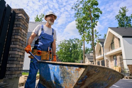 Foto de Alegre trabajador de la construcción con sombrero duro y guantes de goma empujando carretilla hacia adelante entre las casas inacabadas - Imagen libre de derechos