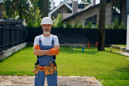Téléchargez les photos : Travailleur de la construction calme en casque dur et gants en caoutchouc posant pour caméra dans un quartier clôturé avec des maisons résidentielles sur fond - en image libre de droit