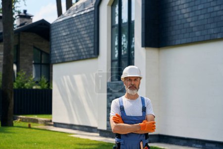 Photo for Waist-up portrait of builder in rubber gloves and safety helmet posing for camera against fenced modern private residential house - Royalty Free Image