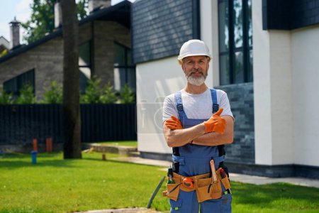 Photo for Smiling construction worker in hard hat and rubber gloves giving thumbs-up while posing for camera against fenced modern cottage - Royalty Free Image