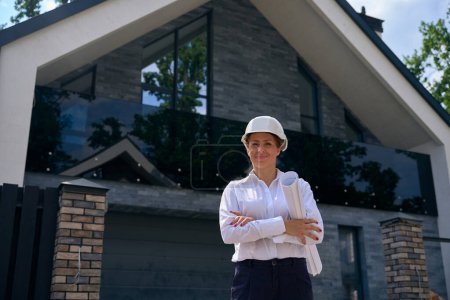 Photo for Smiling female site manager with rolled-up architectural drawings posing for camera against newly built private house during site inspection - Royalty Free Image