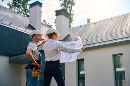Foto de Contratista alegre señalando el techo de la nueva casa mientras mira los dibujos arquitectónicos en manos del gerente de construcción - Imagen libre de derechos