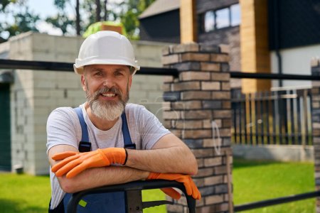 Photo for Waist-up portrait of contented builder leaning on platform trolley handle while standing near fenced building plot - Royalty Free Image