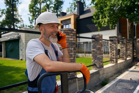 Photo for Waist-up portrait of smiling building contractor in hard hat leaning on platform trolley handle during phone conversation near fenced plot - Royalty Free Image
